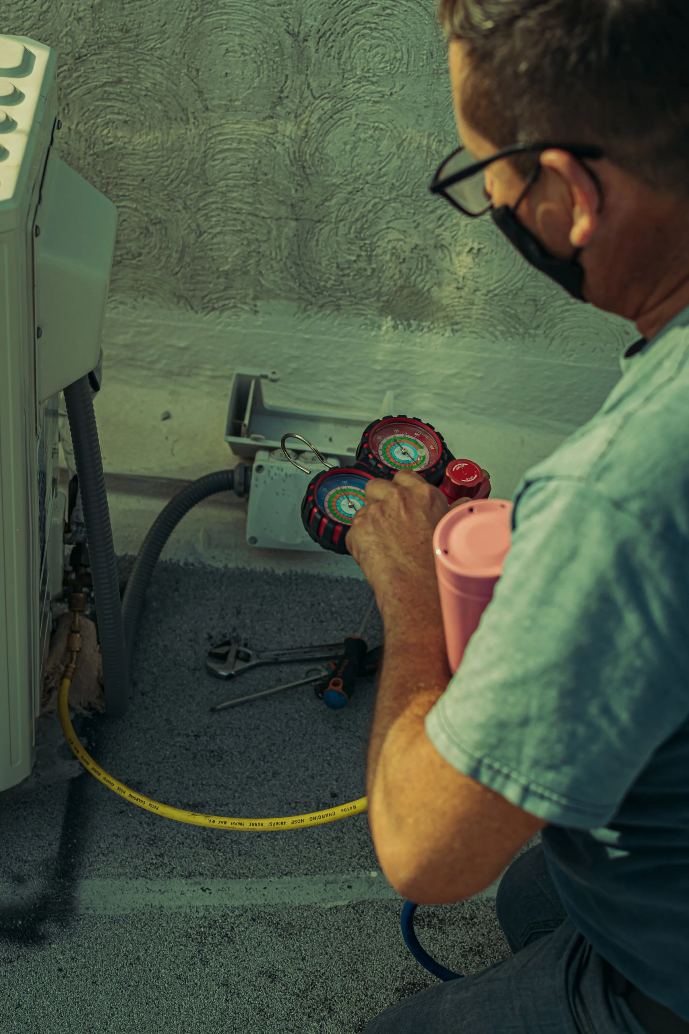 HVAC technician using a refrigerant gauge manifold to service an outdoor air conditioning unit, tools laid out on the ground beside him